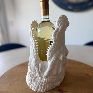May include: A white alligator-shaped wine bottle holder cradles a bottle of white wine. The holder is made of a textured material, with the alligator's jaws open to hold the bottle. The wine bottle has a gold-colored top.