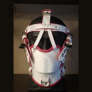 May include: A white and pink leather head harness with metal fixtures. The design incorporates straps, buckles, and a central face covering. It features a neck strap and a lock and key detail.