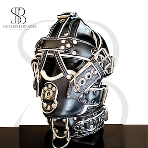 Puede incluir: Una capucha de cuero negro con costuras blancas y remaches plateados. La capucha presenta múltiples correas, hebillas y un anillo de metal en la base. El texto "SAIM ENTERPRISES LEATHER ART" es visible.