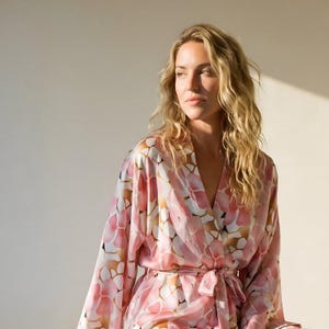 May include: A long-sleeved robe featuring a repeating pattern of soft pink, white, and tan organic shapes. The garment has a V-neckline, wide sleeves, and a matching fabric tie belt at the waist, crafted from a smooth, lightweight material.