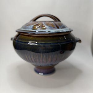 May include: A ceramic lidded serving bowl with a handle. The bowl is a deep blue with brown and purple accents. The lid has a handle and a textured surface. The bowl has a round shape and sits on a small base.