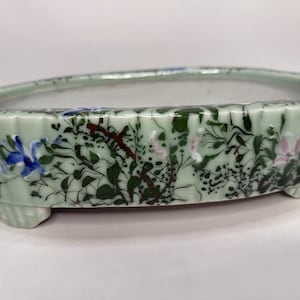 以下が含まれることがあります： 花柄のライトグリーンのセラミック盆栽鉢。 楕円形の鉢はスカラップエッジで、青、ピンク、緑の花と葉で飾られています。 鉢は3つの小さな足の上にあります。