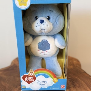 Może przedstawiać: Niebieska pluszowa zabawka Grumpy Bear z edycji kolekcjonerskiej na 20-lecie Care Bears, zapakowana w niebieskie kartonowe pudełko z przezroczystym okienkiem. Miś ma na białym brzuchu ciemnoniebieski symbol chmury deszczowej i nos w kształcie serca.