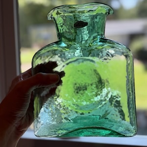 Pode incluir: Uma bolsa de água quente de vidro verde-claro translúcido com uma superfície texturizada e martelada. A garrafa apresenta uma abertura de gargalo larga e alargada e uma pequena etiqueta oval escura perto da borda superior com texto visível.