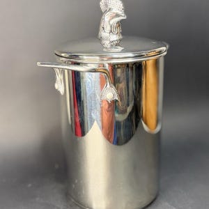 Op de afbeelding: Zilveren ijsemmer met deksel en decoratieve handgreep in de vorm van een haan. De emmer heeft een cilindrische vorm en handvatten aan de zijkanten. De zilveren deksel past op de emmer.