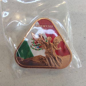 May include: A triangular copper-toned metal coin featuring a relief of the Winged Victory of Angel of Independence statue. The background displays the Mexican flag in red, white, and green enamel. The text LIBERTAD and 2024 are embossed at the top.