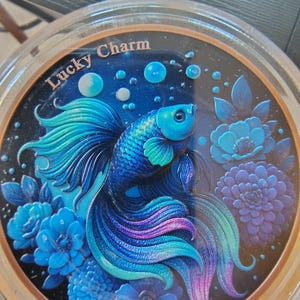 May include: A circular decorative object featuring a vibrant blue and purple betta fish swimming among stylized blue flowers and bubbles. The text Lucky Charm is printed in gold lettering at the top of the dark background within a clear casing.