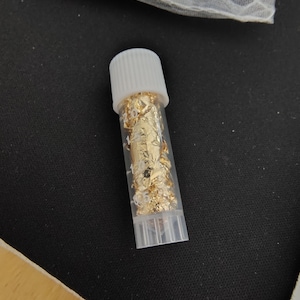 May include: A small, clear plastic vial filled with gold leaf flakes, capped with a white lid. The vial sits on a dark surface, with a partially visible mesh bag containing more gold leaf in the background.