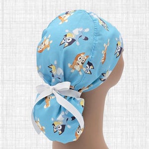 May include: A light blue scrub cap featuring a repeating pattern of cartoon dogs in shades of blue, orange, and white. The cap has a gathered back section secured with a white ribbon tied into a bow, designed to accommodate long hair securely.