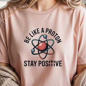 May include: A light pink t-shirt with the text "BE LIKE A PROTON STAY POSITIVE" in black. The shirt features a graphic of an atom with a red center and blue and black orbiting lines. The shirt is short-sleeved.