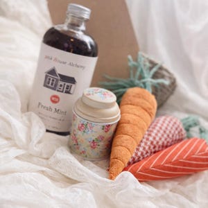 May include: A collection of decorative items including a bottle of Pink House Alchemy Fresh Mint, a floral-patterned tin, and plush carrot and leaf-shaped ornaments. The items are arranged on a white, textured surface.