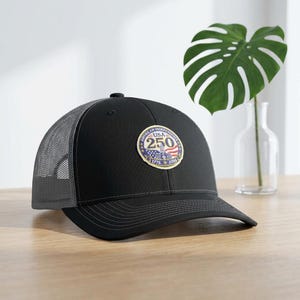 Peut inclure: Casquette de camionneur noire avec un dos en filet et une visière incurvée. L'avant présente un écusson circulaire avec le texte "AMERICAN INDEPENDENCE USA 250" et les dates 1776 et 2026, en rouge, blanc et bleu.