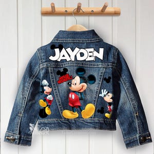 Puede incluir: Chaqueta vaquera azul con dibujos de Mickey Mouse y el nombre "JAYDEN" en letras blancas. La parte trasera de la chaqueta tiene la inscripción "MOUSE-KETASTIC!". Cuelga de una percha de madera.