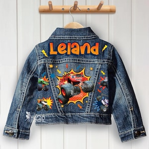 May include: A blue denim jacket with a colorful monster truck graphic on the back. The name "Leland" is printed in orange and yellow across the top. The jacket has button closures and a classic collar.