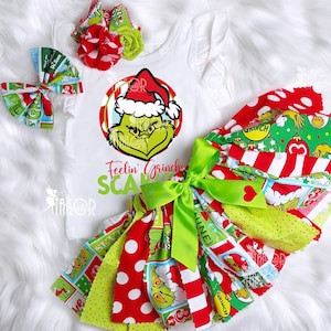 May include: A white Grinch-themed outfit with a matching bow and hair accessories. The top features a Grinch graphic and the words "Feelin' Grinchy". The skirt is a mix of red, green, and white fabrics. The bows and skirt have a festive Christmas theme.
