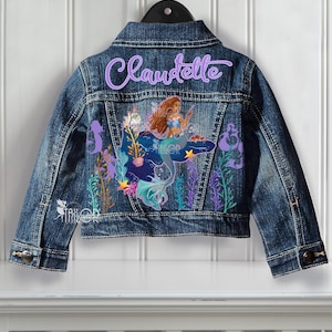 May include: A blue denim jacket featuring a mermaid design and the name "Claudette" embroidered on the back. The mermaid has brown hair and a blue tail, with colourful aquatic plants surrounding her.
