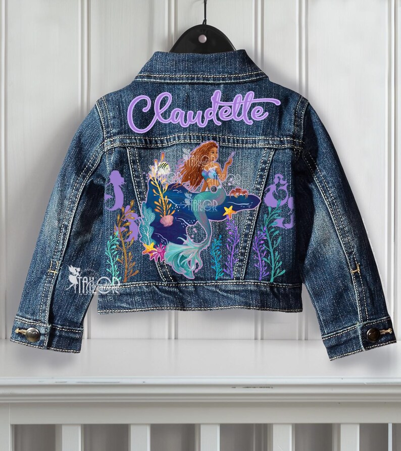 May include: A blue denim jacket featuring a mermaid design and the name "Claudette" embroidered on the back. The mermaid has brown hair and a blue tail, with colourful aquatic plants surrounding her.