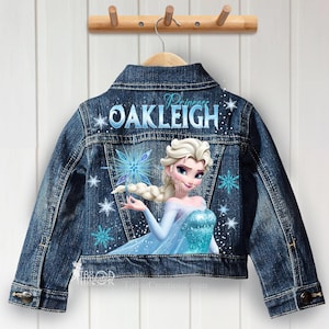 May include: A denim jacket with a blue and white illustration of a princess. The jacket features the name "Oakleigh" in blue script and snowflake accents. The jacket is hanging on a wooden hanger.