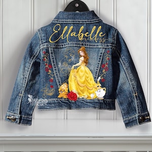 Puede incluir: Chaqueta vaquera azul con un diseño que presenta un vestido amarillo, el texto "Ellabelle Princess" y otros personajes. La chaqueta tiene una parte delantera abotonada y está colgada de una percha negra.