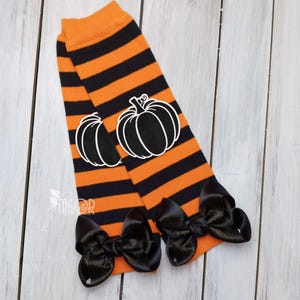 Puede incluir: Calentadores de piernas a rayas naranjas y negras con lazos negros en la parte inferior y aplicaciones de calabaza con contorno blanco. Estos calentadores de piernas con temática de Halloween son perfectos para un atuendo festivo.