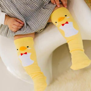 May include: Yellow knee-high socks with a cartoon chick design. The socks feature a chick face with a red bow tie and a white body shape. The socks are worn by a child.