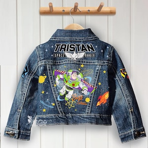 Puede incluir: Chaqueta vaquera azul con un diseño de Buzz Lightyear y el nombre "Tristan" en la espalda. La chaqueta presenta parches coloridos e ilustraciones de estrellas, planetas y un cohete. La chaqueta está colgada en una percha de madera.