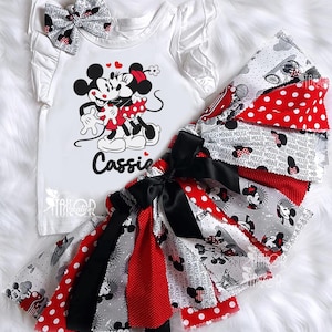 May include: A white t-shirt with ruffled sleeves and a matching bow, featuring a Minnie and Mickey Mouse graphic. The set includes a red, black, and white skirt with Minnie Mouse patterns and polka dots. The name "Cassie" is printed on the shirt.