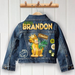May include: A blue denim jacket with a Simba design. The back of the jacket features the name "BRANDON" in gold lettering above a Simba graphic. Additional patches include paw prints and a Simba label.