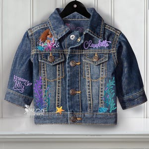 May include: A blue denim jacket with embroidered details. The jacket features a mermaid design, the name "Claudette" in purple script, and the phrase "dream BIG". Decorative elements include a starfish and seaweed.