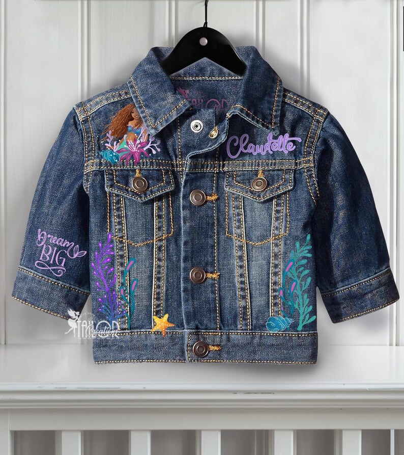 May include: A blue denim jacket with embroidered details. The jacket features a mermaid design, the name "Claudette" in purple script, and the phrase "dream BIG". Decorative elements include a starfish and seaweed.