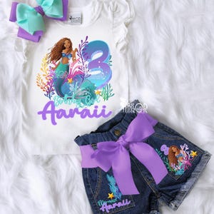 May include: A white t-shirt and denim shorts set with a mermaid theme. The shirt features a mermaid graphic and the number 3. The shorts have a purple bow and mermaid details. A matching bow completes the set.