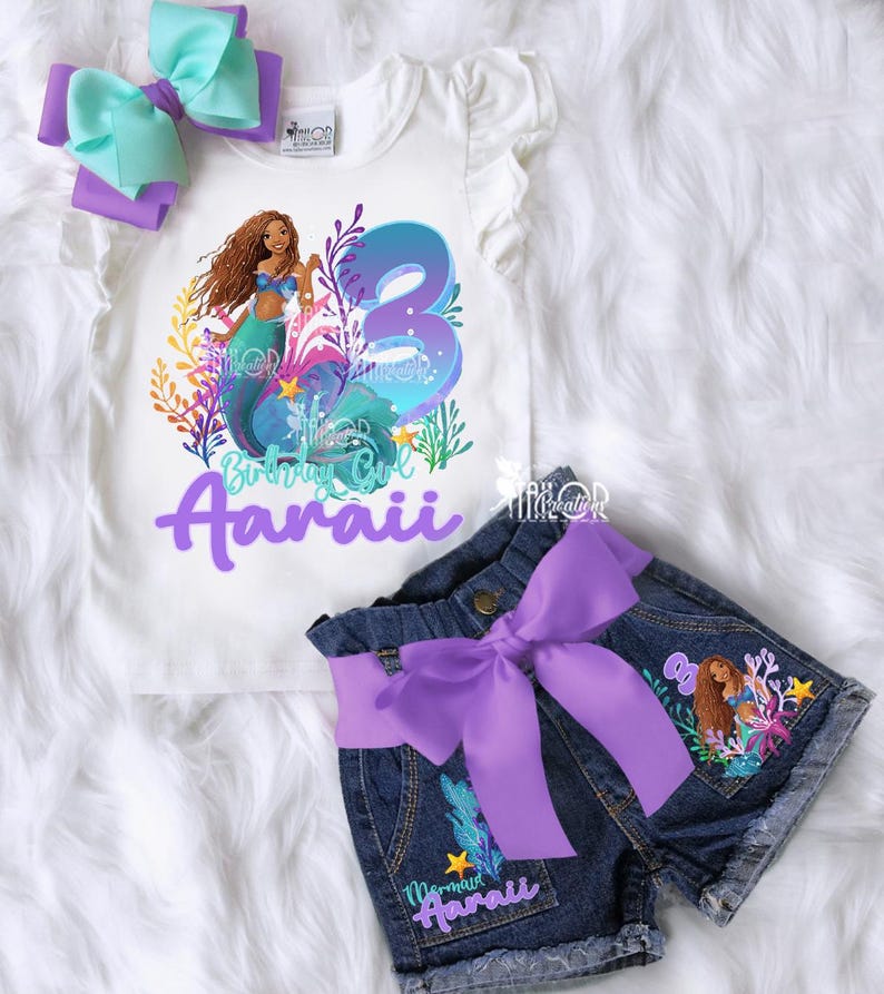 May include: A white t-shirt and denim shorts set with a mermaid theme. The shirt features a mermaid graphic and the number 3. The shorts have a purple bow and mermaid details. A matching bow completes the set.