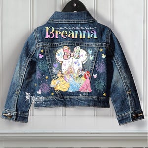 Może przedstawiać: Granatowa kurtka dżinsowa z kolorowym wzorem. Tył kurtki zdobi imię "Breanna" w tęczowych literach, głowa Myszki Minnie, księżniczki Disneya, fajerwerki i gwiazdy. Kurtka wisi na czarnym wieszaku.