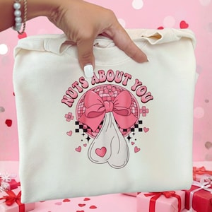 以下が含まれることがあります： ピンク色の文字で「Nuts About You」と書かれたグラフィックが特徴の、クリーム色のスウェットシャツ。ピンクのリボン、ディスコボール、ハート型のオブジェクトが描かれています。スウェットシャツは手で持たれています。