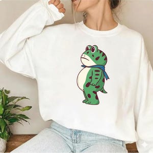 Op de afbeelding: Witte sweatshirt met een cartoon kikker ontwerp. De kikker is groen met donkerrode vlekken, een blauwe sjaal en een neutrale uitdrukking. Het ontwerp is gedrukt op de voorkant van de witte sweatshirt. De kikker heeft een witte buik.