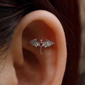 May include: A silver dragon-shaped earring with a red gemstone center. The dragon has detailed wings and a curved tail. The earring is shown on an ear, highlighting its size and design.