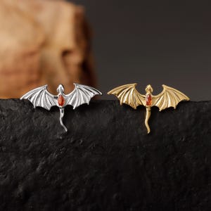 May include: Two dragon-shaped brooches. One is silver-toned, the other gold-toned. Each has outstretched wings and a red gemstone in the center. The brooches are displayed on a black surface, with a blurred background.