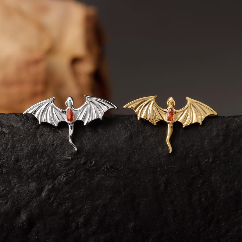 May include: Two dragon-shaped brooches. One is silver-toned, the other gold-toned. Each has outstretched wings and a red gemstone in the center. The brooches are displayed on a black surface, with a blurred background.