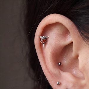 May include: A silver-toned ear piercing stud featuring a central heart-shaped red gemstone, flanked by wing-like accents and a pointed base. The jewelry is positioned in the upper cartilage area, accompanied by two small silver ball studs below.