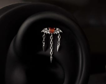 May include: A silver earring with a bat motif, incorporating a red heart-shaped gemstone at its centre. The bat's wings are outstretched, and three chains with pointed tips hang beneath. The earring is designed for ear placement.