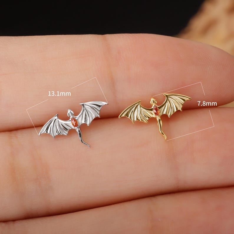 May include: Two dragon-shaped earrings. One is silver-colored, the other is gold-colored. Each dragon has outstretched wings and a red gemstone. The silver dragon measures 0.5 inches long. The gold dragon measures 0.3 inches long.