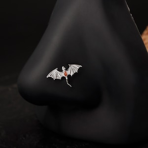 May include: A silver nose stud in the shape of a dragon with outstretched wings and a red gemstone. The dragon is detailed with textured wings and a slender tail. The stud is set against a black background.