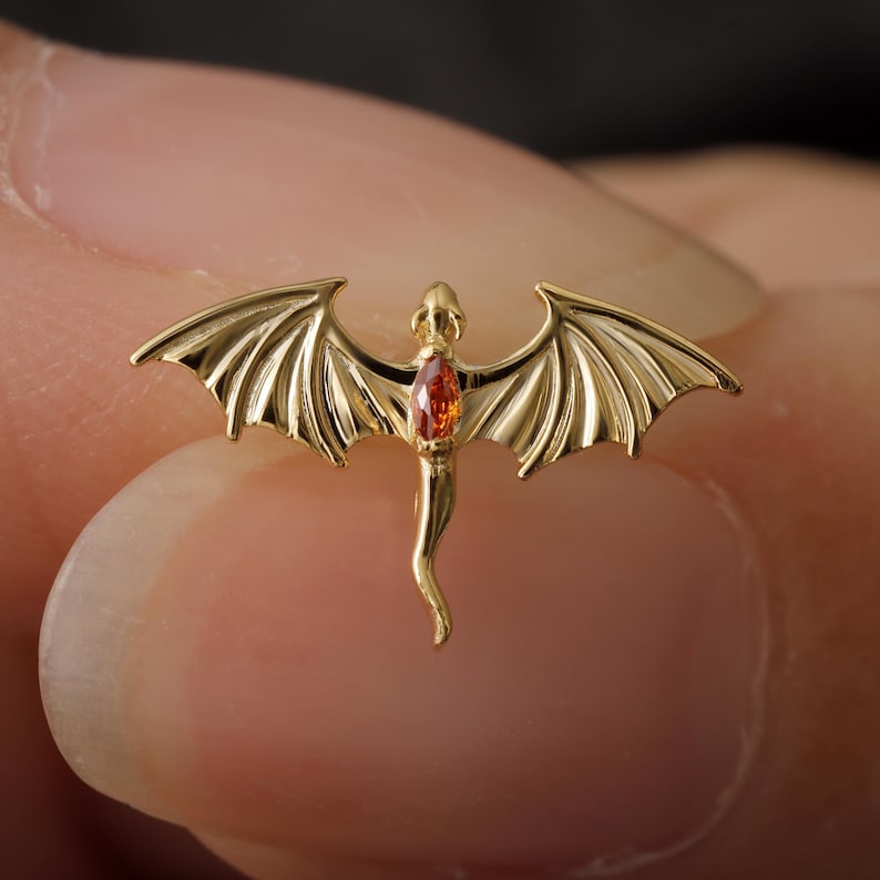 May include: A gold-colored dragon pendant with outstretched wings and a gemstone center. The dragon has detailed wing and body features. The gemstone is a vibrant orange color. The pendant is likely jewelry.