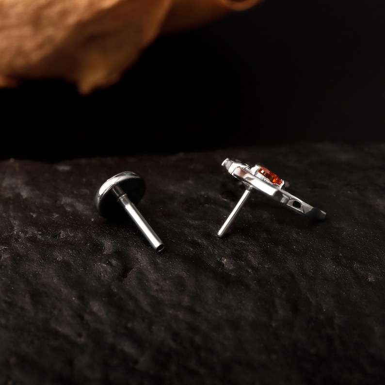 May include: Two pieces of jewelry. One is a silver-colored stud with a dark, round top. The other is a silver-colored stud with a small, decorative design and an orange-colored stone. Both have a post for piercing.