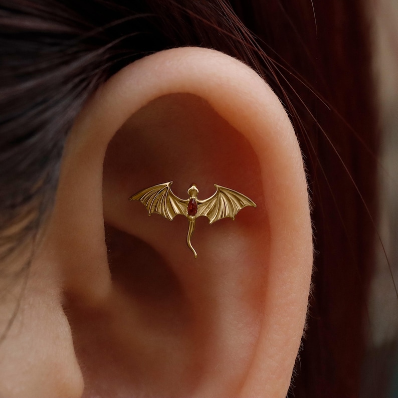 May include: A gold-colored dragon-shaped earring with a red gemstone center. The dragon has outstretched wings and a curved tail. The earring is designed to be worn on the ear.