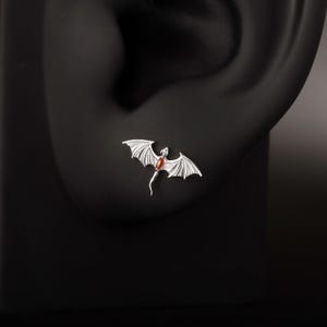 May include: A silver dragon earring with a red gemstone center. The dragon has detailed wings and a slender body. The earring is displayed on a black background, showcasing the jewelry's design.
