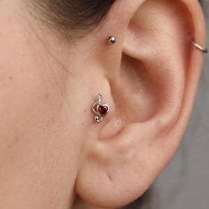 May include: A silver-toned ear piercing stud shaped like a musical treble clef with a small, deep red heart-shaped gemstone set in the center. The jewelry is positioned in the tragus area of the ear, accompanied by a simple silver ball stud above.