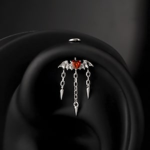 Puede incluir: Joya para la oreja plateada con un diseño de ala de murciélago y una piedra central roja en forma de corazón. Tres cadenas con puntas cuelgan por debajo. La joya está colocada sobre un fondo oscuro.