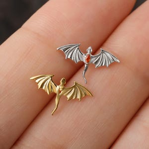 May include: Two dragon-shaped jewelry pieces. One is silver with a red gem, the other is gold. The dragons have outstretched wings and detailed bodies. The jewelry is small and delicate, suitable for earrings or pendants.