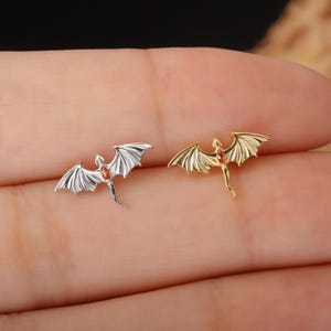 May include: Two small dragon-shaped earrings. One is silver-colored, the other is gold-colored. Both have outstretched wings and a small red gem in the center of the body. The earrings are displayed on a light-colored background.